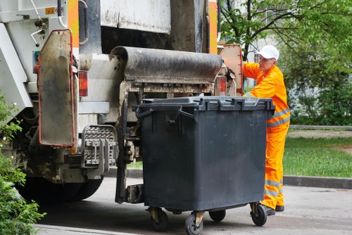 A worker dressed in a high-visibility orange uniform and white cap operates a large commercial rubbish compactor mounted on the side of a waste collection vehicle. The worker stands on a paved surface next to a black wheeled rubbish bin, which is positioned in front of the compactor's loading mechanism. The bin is made of plastic with a textured surface and a fitted lid. The waste collection vehicle, with its metal body and hydraulic components, is partially visible, showing the conveyor and loading area used for compacting rubbish. In the background, there are green trees and a grassy verge, indicating an outdoor setting, possibly near residential or commercial premises within the Chertsey area. The lighting is natural and suggests daytime with overcast skies, emphasizing the work environment in waste management services provided by Commercial Waste Chertsey for commercial rubbish removal or general waste disposal needs in the local postcode area.