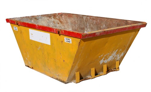A large industrial skip situated on an outdoor paved area, painted in bright yellow with a red reinforced rim along the top edge. The skip's interior, visible from the top, shows accumulated waste material with a rusted, weathered metal surface. The exterior surface features some dirt and minor paint chipping, typical of frequent use in waste management. The skip has a rectangular shape with slightly tapered sides, positioned on a flat ground with no surrounding objects visible. In the background, a neutral outdoor setting is evident, suggesting it is part of a commercial waste removal operation, likely managed by a company such as Commercial Waste Chertsey. This type of heavy-duty skip is used for collecting bulk rubbish and debris, and its placement on a driveway or yard aligns with practical waste disposal needs within the local area, possibly near Chertsey or nearby towns. The scene reflects a professional waste collection setup dedicated to commercial rubbish removal services.