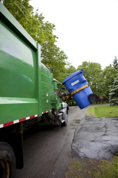 A green commercial waste collection truck is parked on a driveway or street, with a large metal hopper or storage compartment at the rear. The container is painted in a solid, bright green finish and is designed for holding refuse before removal. Adjacent to the truck, a blue wheeled rubbish bin is tilted at an angle, with its black wheels visible on the pavement, indicating it is in the process of being emptied or collected. The bin has a matching blue lid, secured with a yellow strap or handle. The surrounding environment features a paved surface that appears to be part of a residential or semi-urban area, with some cracked or uneven pavement visible. In the background, there are lush, green-leaved trees and a cloudy sky, suggesting an overcast day. The scene depicts a typical rubbish removal operation, with the truck positioned to facilitate waste collection, conducted by Commercial Waste Chertsey, highlighting their service of rubbish removal within the local area near Chertsey.