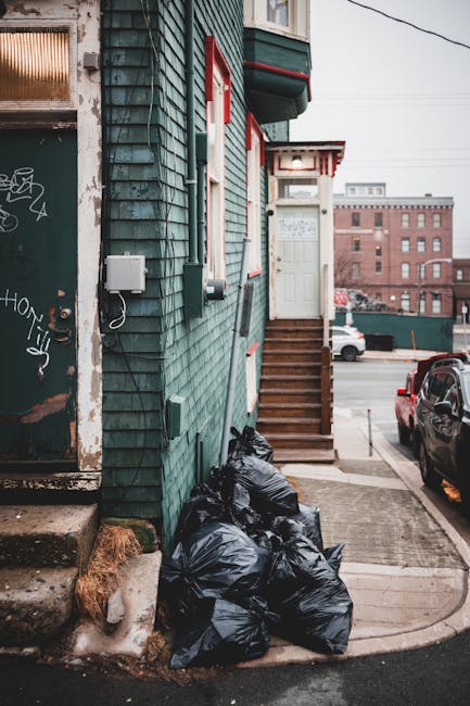 A group of black plastic rubbish bags, filled and tied, are piled at the base of a green wooden building with weathered, shingle-style exterior cladding. The bags are situated on a concrete sidewalk n