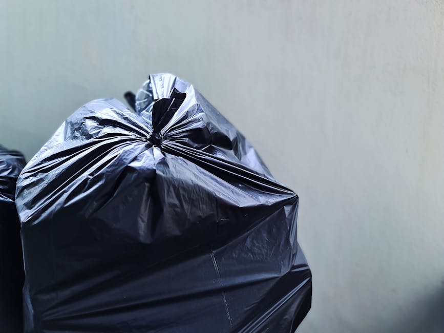 A group of black plastic rubbish bags, filled and tied, are piled at the base of a green wooden building with weathered, shingle-style exterior cladding. The bags are situated on a concrete sidewalk n